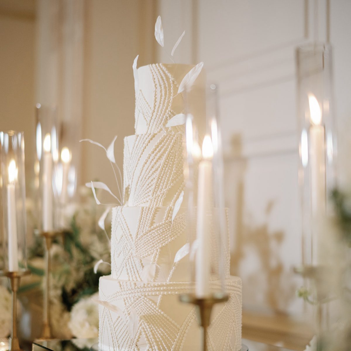 Four-tier white wedding cake with beaded piping and white feathers among candles