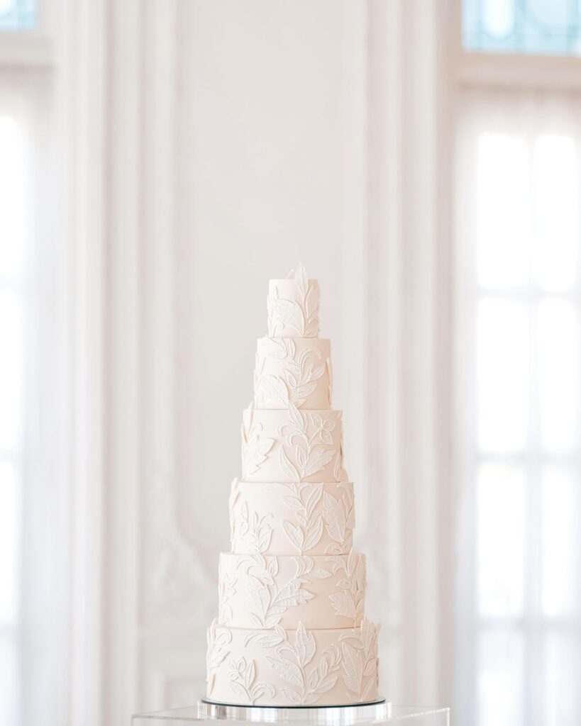 Six-tier ivory wedding cake with sugar leaf appliqués in a bright white salon