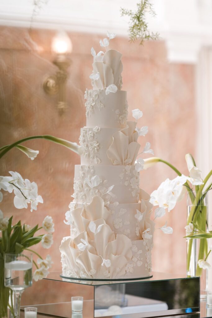 Six-tier ivory wedding cake with sculpted sugar petals and delicate flowers, beside orchids and calla lilies